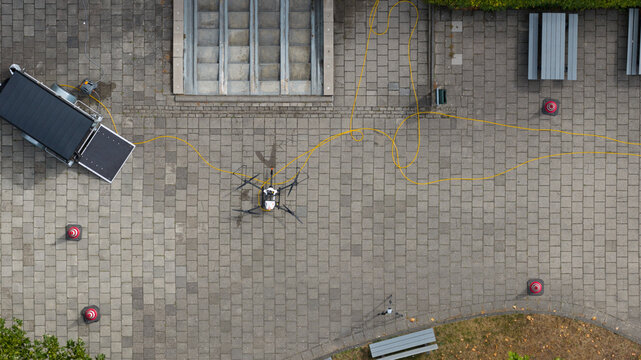 Aerial view of a drone connected by a yellow cable to a trailer on a paved plaza with benches and traffic cones in Amersfoort, Utrecht, Netherlands.