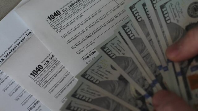 Close-up shot of a fan of hundred-dollar bills held over official US Individual Income Tax Return Form 1040 documents. Person holding cash money for paying tax duties 