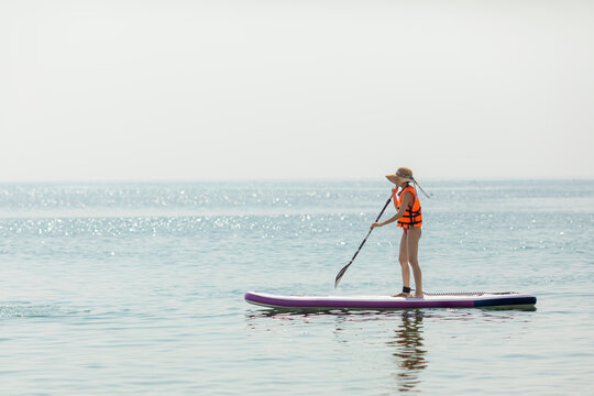 Young girl wearing an orange life jacket stands on a purple stand-up paddleboard, using a paddle to navigate calm ocean waters, with a clear sky and distant horizon visible in the background