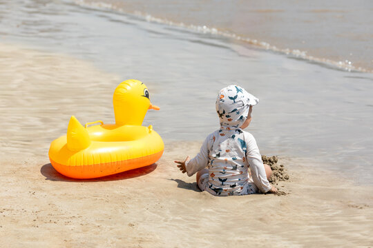 Rear view child in a sun hat and swimwear sits on the sandy beach, playing with sand while an inflatable yellow duck floats nearby, with gentle waves and a calm sea in the background