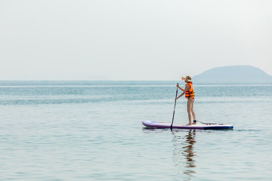 Young girl wearing an orange life jacket stands on a purple stand-up paddleboard, using a paddle to navigate calm ocean waters, with a clear sky and distant horizon visible in the background