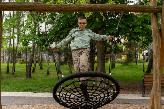 School age boy balancing on large suspended rope swing at outdoor playground in green park. Concept of active childhood, physical development, coordination, balance training and healthy lifestyle. 