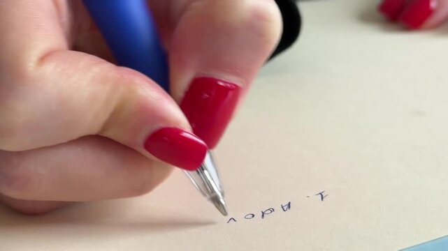 A hand is writing with a blue pen on a beige surface.