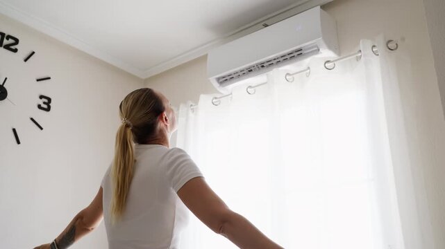 Relaxed woman standing beside air conditioner with arms raised, enjoying refreshing cool air at home during a hot summer day and experiencing comfort after escaping the heat