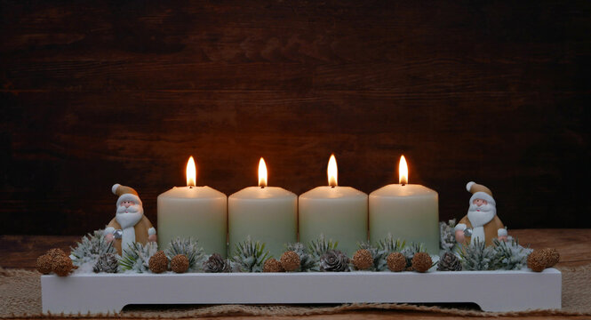 4th Sunday of Advent: An Advent wreath with four green candles, all of which are lit, featuring pinecones, small fir branches, and two Santa Claus figures against a wooden background with space for te