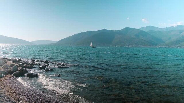 Beautiful Tivat beach with gentle waves on the clear blue sea, boats floating offshore and majestic green mountains rising in the distance, creating a peaceful summer coastal scene.