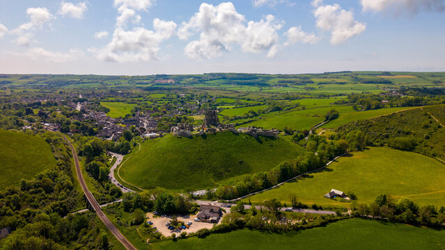 Aerial view of Corfe Castle ruins on a green hill surrounded by the rolling Purbeck countryside, lush meadows, and the historic village, Corfe Castle, England, United Kingdom.