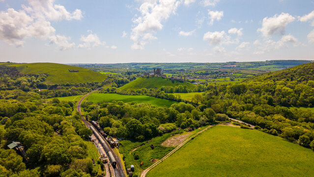 Aerial view of Corfe Castle ruins set amidst the lush green Purbeck Countryside with a heritage steam train at the station in Corfe Castle, England, United Kingdom.