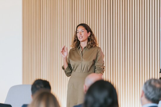 Woman presenting to a seated audience, wearing a muted olive shirt dress against a slatted timber wall for business seminar and conference marketing