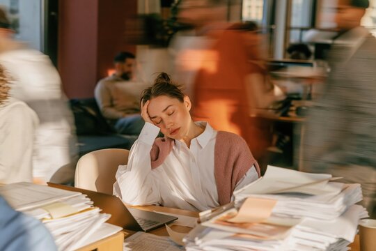 Overwhelmed woman sitting at a desk with eyes closed, feeling exhausted in a busy office with motion blur for workplace stress and employee wellbeing