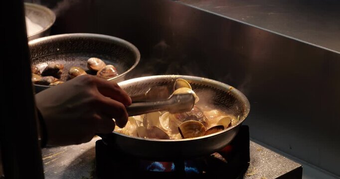 Close up side view of street vendor preparing fresh clams in rich, bubbling sauce at Asian night market stall. cook uses metal tongs to lift and toss seafood shells within pan to stir them thoroughly