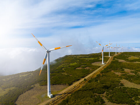 Aerial view of wind turbines standing on a green ridge with a dirt road and clouds in the background in Madeira, Castelo Branco, Portugal.