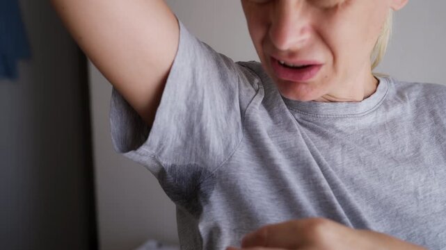 Woman showing a visible sweat stain on her gray T-shirt with a disgusted expression, illustrating excessive underarm sweating, body odor and personal hygiene concerns.