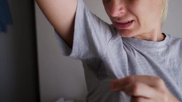 Woman examining a visible underarm sweat stain on her gray T-shirt, illustrating concerns about excessive perspiration, odor control and the need for effective deodorant protection.