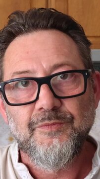 Close-up of a thoughtful mature man with glasses and a beard, looking and blinking. He appears to be speaking, conveying a serious or contemplative expression while engaging in conversation.