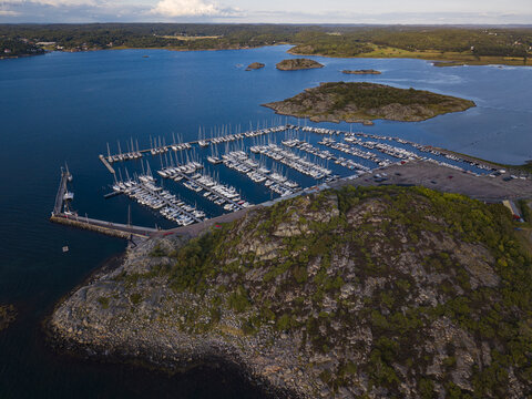 Aerial view of Sando marina with numerous sailboats docked in the harbor, surrounded by rocky coastline, lush green islands, and deep blue sea in Vallda, Hallands lan, Sweden.