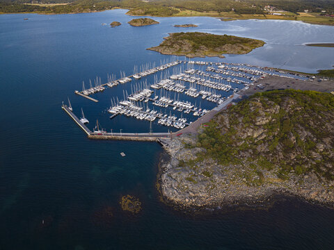 Aerial view of Sando marina with numerous sailboats docked in rows, surrounded by rocky coastline, calm blue water, and green landscape in Vallda, Hallands lan, Sweden.