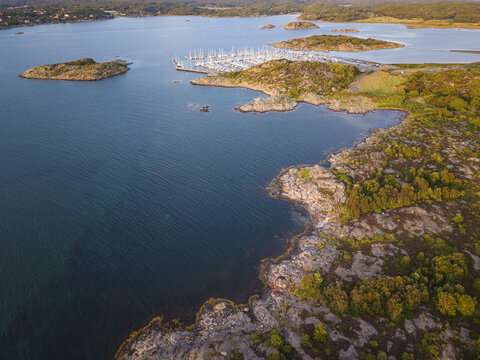 Aerial view of Sando marina with sailboats docked along the rocky coastline surrounded by calm blue water and lush greenery in Vallda, Hallands lan, Sweden.
