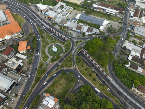 Aerial view of the Statue of Dewa Ruci situated at a busy traffic junction surrounded by urban roads, lush greenery, and buildings in Kecamatan Kuta, Bali, Indonesia.