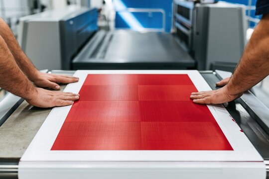 Hands pressing red proof sheets on a printing press delivery table, featuring wet ink sheen and industrial textures for commercial prepress services