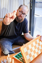 Casual man arranging chess set. Relaxed man demonstrates chessboard arrangement by sunlit window. White bearded man casually presenting chess pieces with sunny backdrop and friendly expression © anatoliy_gleb