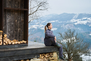 Fototapeta premium Woman sitting on wooden cabin terrace edge, enjoying mountain landscape and quiet nature, reflecting in peaceful moment, embracing slow living and rural lifestyle in winter scenery