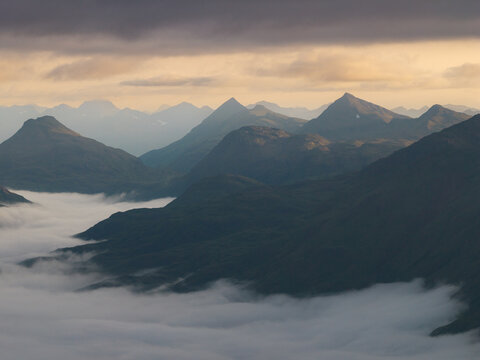 Aerial view of the rugged mountain peaks of Unalaska rising above a thick layer of low-hanging clouds under a soft, golden-hued sky in Unalaska, Alaska, United States.