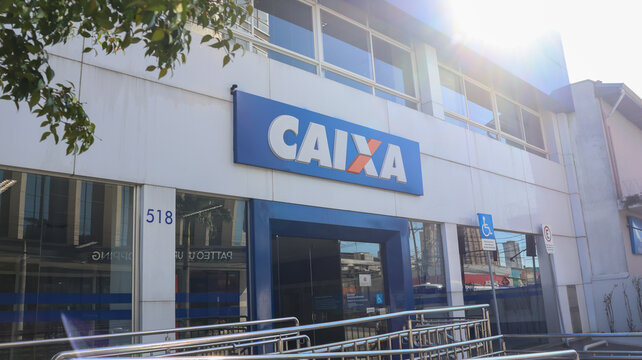 Fachada de ag&ecirc;ncia da Caixa com placa azul, entrada envidra&ccedil;ada, rampa de acesso e arquitetura comercial em &aacute;rea urbana da cidade. Mogi das Cruzes SP 18 agosto 2026