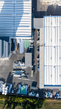 Aerial view of a construction materials manufacturing plant with industrial warehouses, storage areas with pallets and building supplies, and paved loading zones in Milan, Lombardy, Italy.
