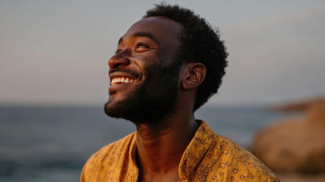 Close-up portrait of a young african-american man with a beard and curly hair. he is wearing a yellow shirt with a floral pattern and is standing on a beach with the ocean in the background.