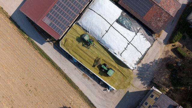 Aerial view of tractors working on silage bunkers at an agricultural farm with solar panels on barn roofs in Peschiera Borromeo, Lombardy, Italy.