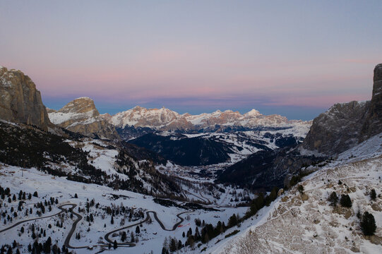 Aerial view of the snow-covered Gardena Pass with its winding mountain road surrounded by the peaks of the Dolomites under a soft twilight sky in Corvara, Trentino-Alto Adige, Italy.