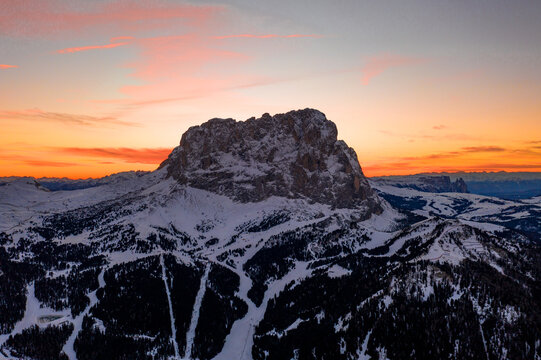 Aerial view of Gardena Pass surrounded by snow-covered peaks and pine forests under a vibrant sunset sky in Corvara, Trentino-Alto Adige, Italy.