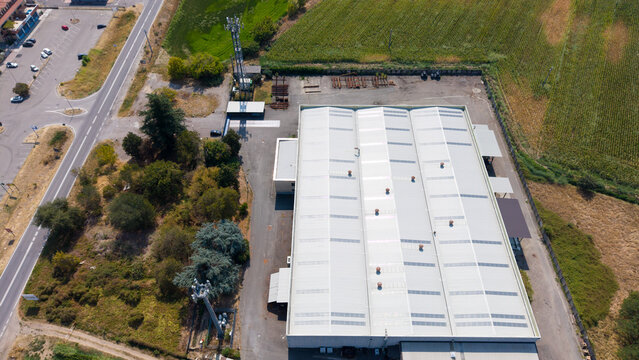 Aerial view of a cold finished steel manufacturing facility with a large white roof, surrounding paved yard, and adjacent agricultural fields in Milan, Lombardy, Italy.
