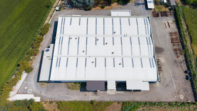 Aerial view of a cold finished steel manufacturing facility with a large white roof, surrounded by paved storage areas and agricultural fields in Milan, Lombardy, Italy.