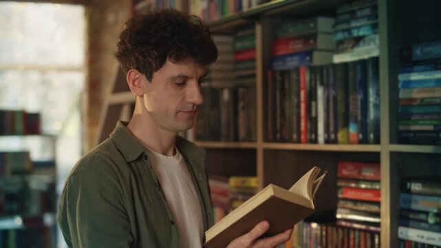 Positive young man holds book and smiles in library, bookworm enjoying hobby