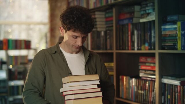 Smiling young male librarian holds stack of books in library, enjoying his job