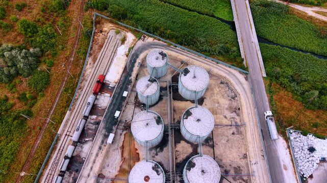 Storage tanks and trucks at a rural industrial site near a road. Storage tanks are arranged in a circular pattern at an industrial site. Trucks are parked nearby on a road.