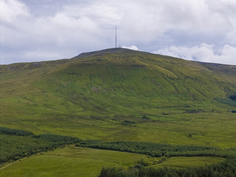 Aerial view of the green slopes of Keeloges mountain topped with a transmission tower, surrounded by rolling fields and patches of forest in County Donegal, Ireland.