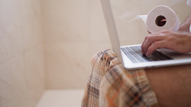 Young man wearing plaid pajama pants sitting on the toilet seat using a laptop computer while holding a roll of toilet paper, representing a funny work-from-home or workaholic concept