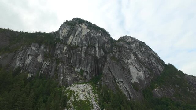 A low angle aerial tracking shot pushes forward over green treetops, gazing up toward the massive, imposing rocky granite rock face of the Stawamus Chief on a cloudy summer day.
