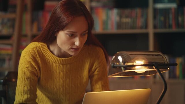 Red-haired female student conducts research in library, typing notes on laptop