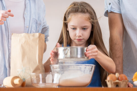Young focused girl child baking in kitchen with family sifting white flour through metal sifter into glass bowl preparing sweet dough for delicious homemade pastry cake dessert food