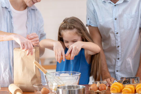 Happy young girl child baking delicious pastry cracking fresh egg into glass bowl with loving mother and father parents together having fun cooking homemade food at wood table