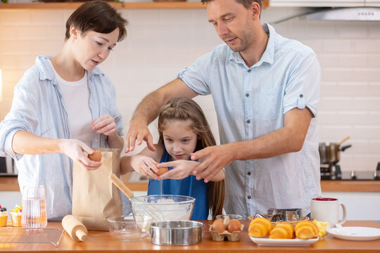 Happy family enjoying baking in kitchen with child. Father and mother assisting daughter with cooking by cracking an egg into bowl, showing focused and joyful expressions together