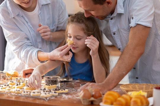 Happy Caucasian family bake together in home kitchen. Mother and father play with child putting dough on girl nose causing joyful emotional expression