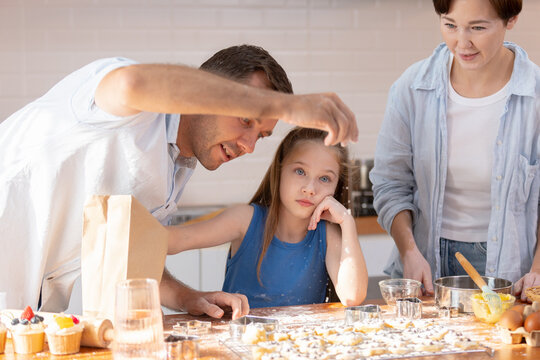 Bored young girl rests head on hand at kitchen table with flour while playful father interacts and caring grandmother watches during family baking activity