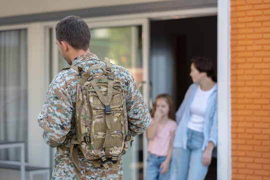 Military soldier camouflage uniform with backpack stands outside house looking wife and daughter doorway feeling deep love and bittersweet separation before deployment or happy homecoming