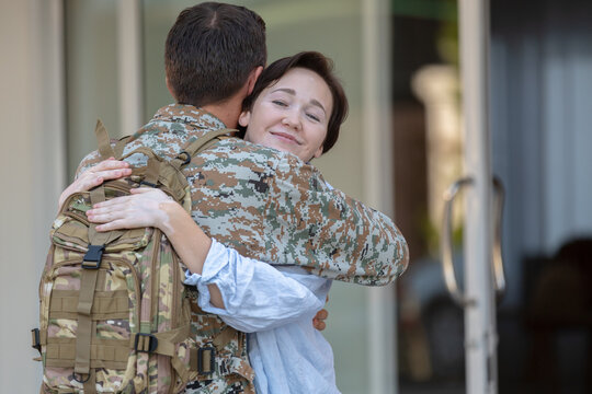 Happy wife feeling deep relief sharing emotional embrace greeting military soldier wearing camouflage uniform returning homecoming holding tactical backpack standing outside door