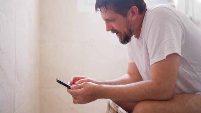 Concentrated man with a beard playing a video game on his mobile phone with great excitement while sitting on the toilet bowl in the bathroom, showing a range of emotions from focus to joy